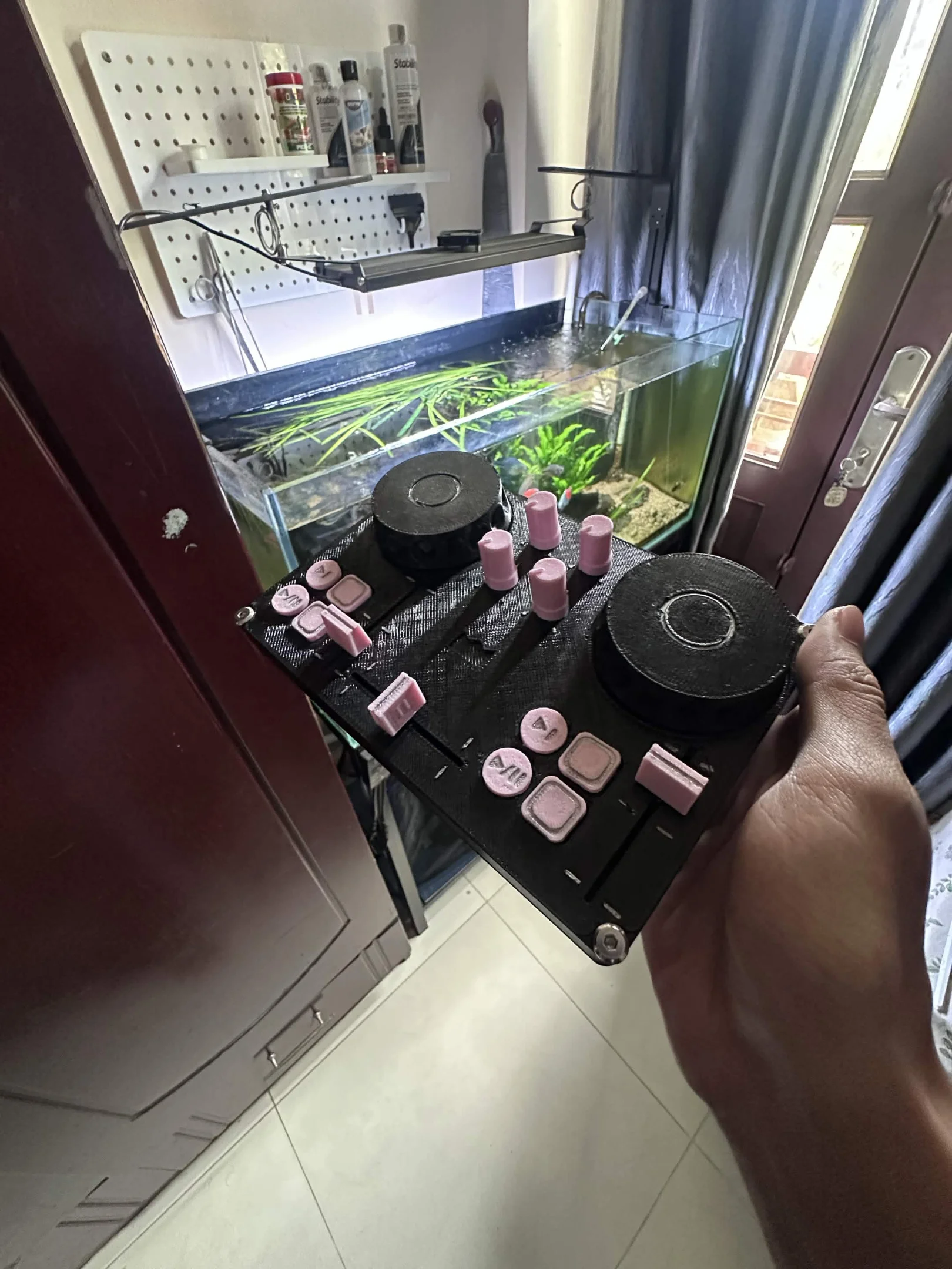 DJC-DIY, a DIY DJ controller printed in black with pink buttons and knobs, held in a hand in front of a planted aquarium