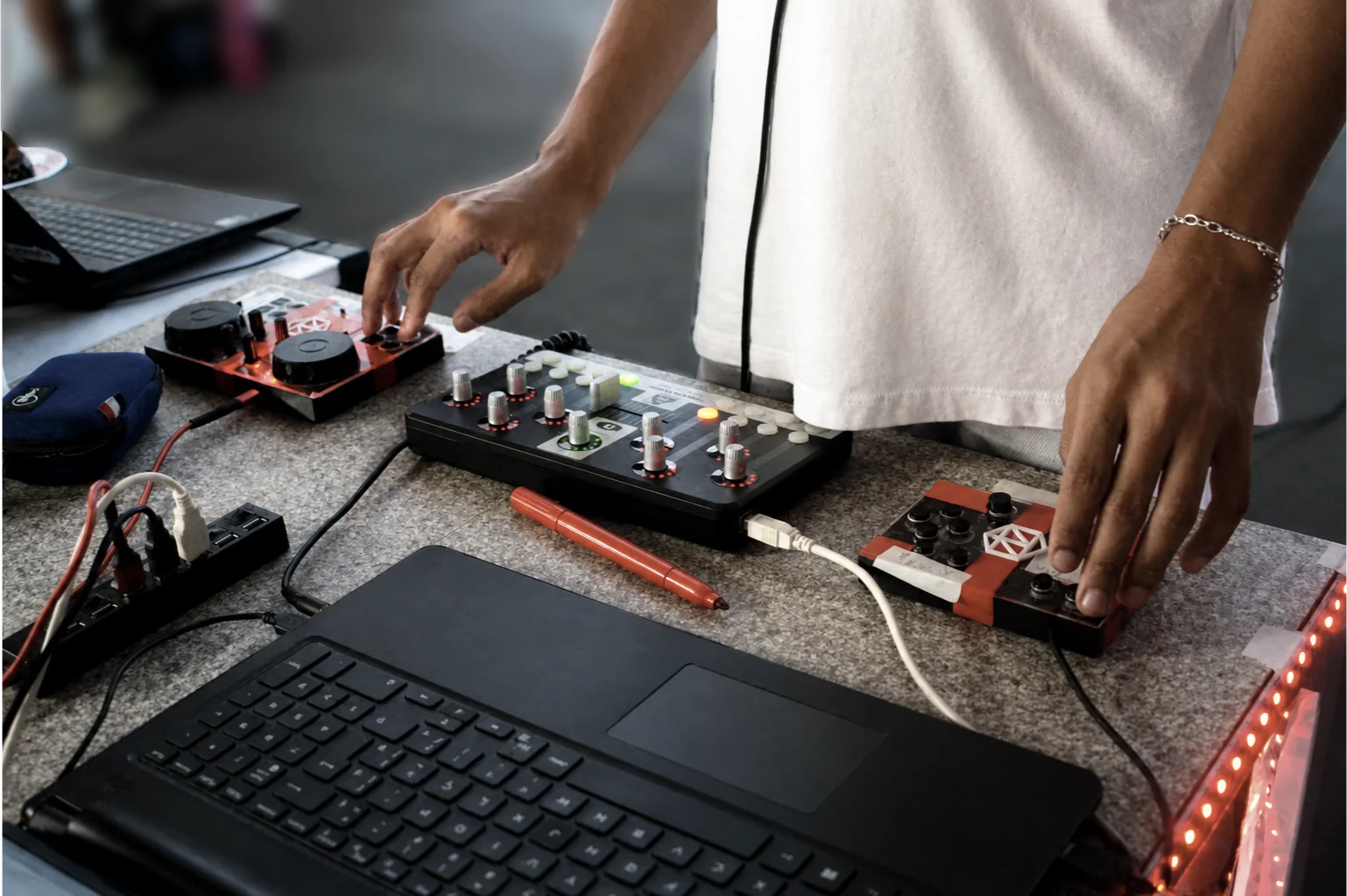 Two DJC-DIY DIY DJ controllers printed in red and white, on a table next to a laptop and a groovebox, with a pair of hands playing on them