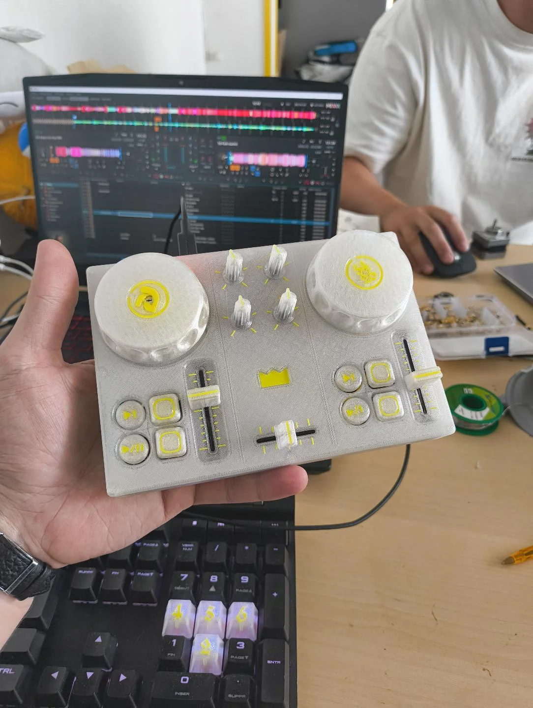 DJC-DIY, a DIY DJ controller printed in white and yellow, held in a hand in front of a monitor running DJ software