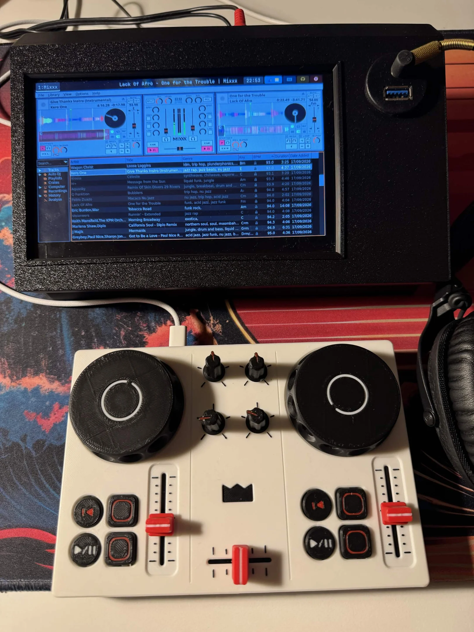 DJC-DIY, a DIY DJ controller printed in white with black jog wheels and red faders, in front of a small screen running Mixxx, with headphones beside it